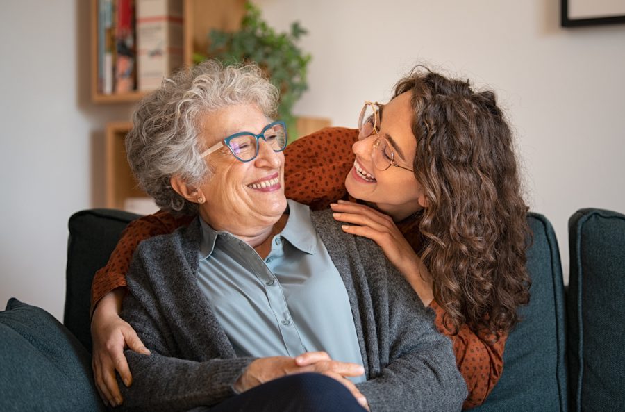 Grandmother and granddaughter laughing and embracing at home Old grandmother and adult granddaughter hugging at home and looking at each other. Happy senior mother and young daughter embracing with love on sofa. Happy young woman hugging from behind grandma with love.