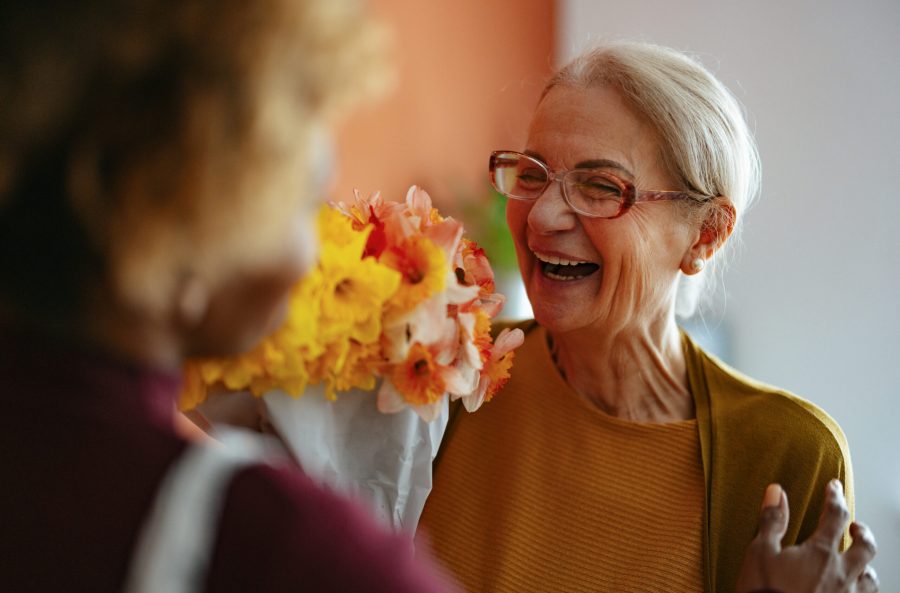 Joyful Senior Woman Receiving Flowers From Caregiver at Home An elderly woman laughing joyfully as she receives a bouquet of flowers from a compassionate caregiver. A touching moment of care and connection in a warm environment.