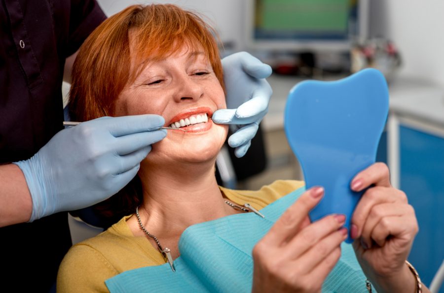 All on 4 Smiling senior woman with new dental implants sitting in the dental office and looking at the mirror